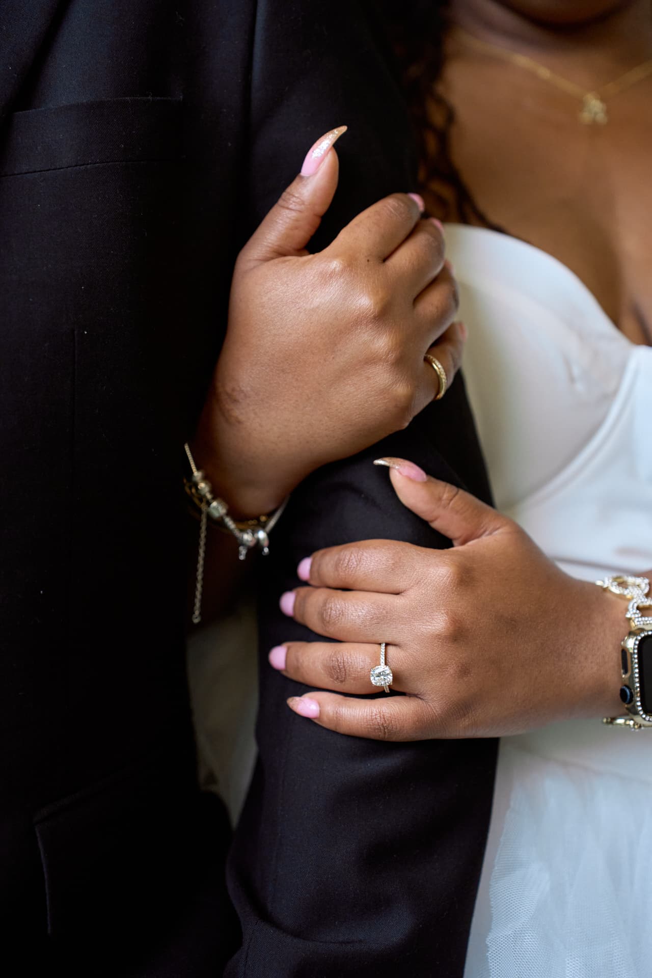 Her hands wrapped around his black suit sleeve, rings and gold jewelry against a white dress